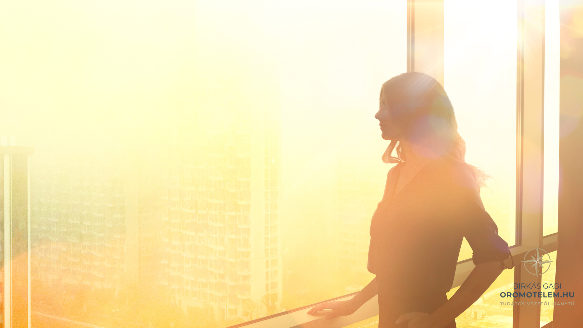 Női vezető áll az ablak előtt napfényben, elgondolkodva, ahogy visszatér a női energiájába és az örömből való teremtésbe.